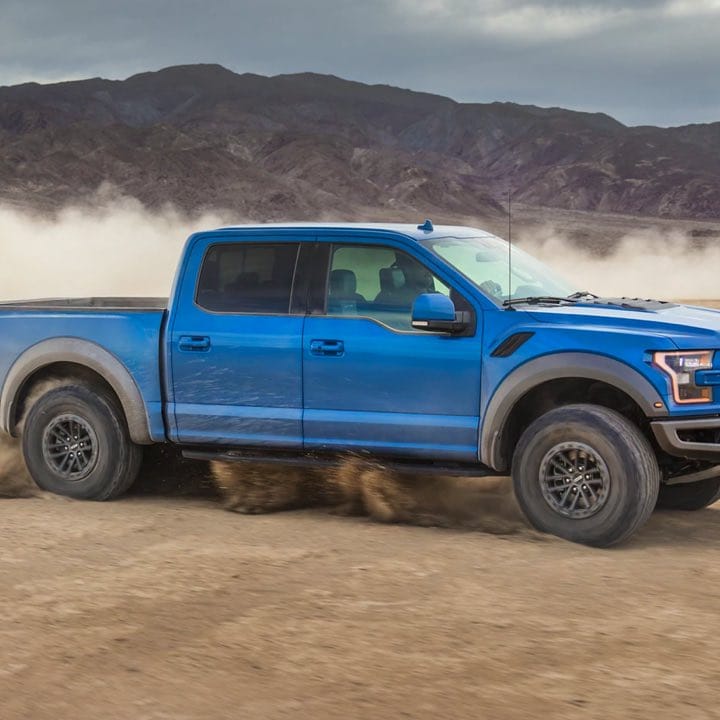 A side photo of a Blue F-150 Raptor driving on a sandy dessert.