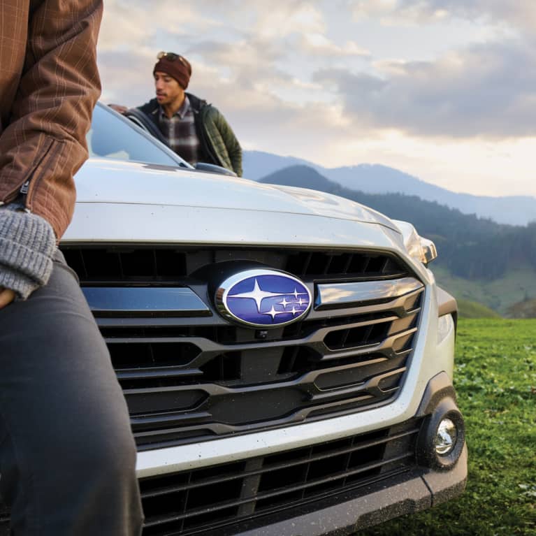 A man and woman are looking at a waterfall, and their blue Subaru is parked on the street behind them.